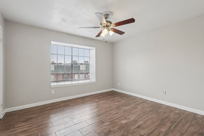 Bright empty room with elegant wood-style flooring, large window, and modern ceiling fan, exuding a fresh and spacious ambiance.