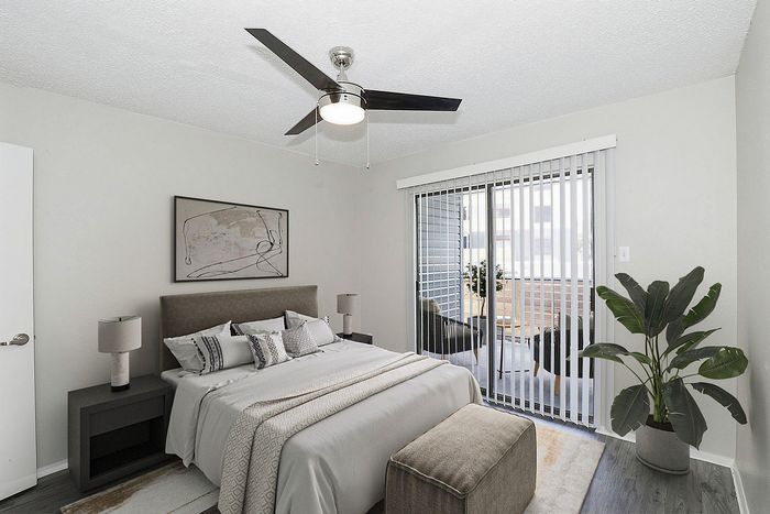 Elegant bedroom with modern decor, featuring a plush bed, stylish ceiling fan, large window with balcony view, and vibrant indoor plant.