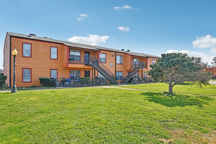 Charming two-story apartment complex with vivid orange exterior, set against a picturesque blue sky. Features inviting outdoor staircases and well-maintained green lawns.