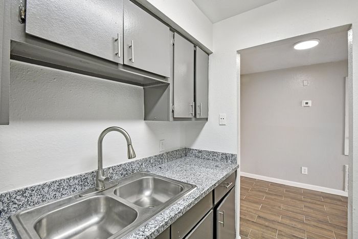 Modern kitchen featuring sleek gray cabinets, stainless steel sink, and stylish granite countertops, leading into an inviting open space with warm wooden flooring.