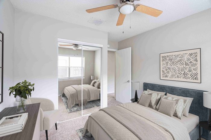 Bright and elegant bedroom with a chic ceiling fan, mirrored closet doors, and a stylishly decorated bed, creating a spacious and inviting ambiance.