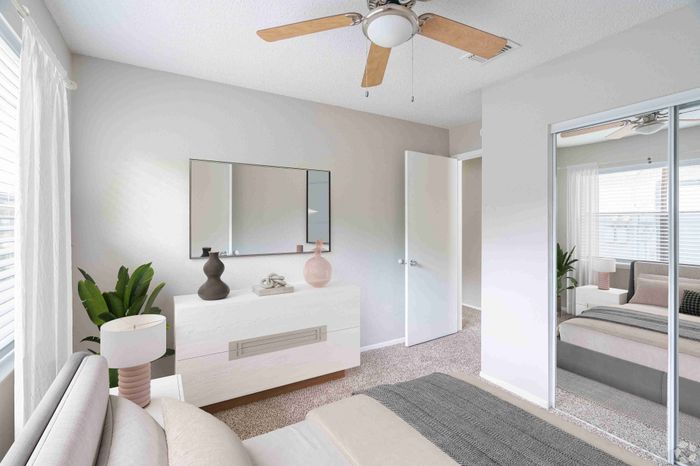 Inviting bedroom with contemporary decor, featuring a stylish white dresser, chic mirror, elegant ceiling fan, and abundant natural light.