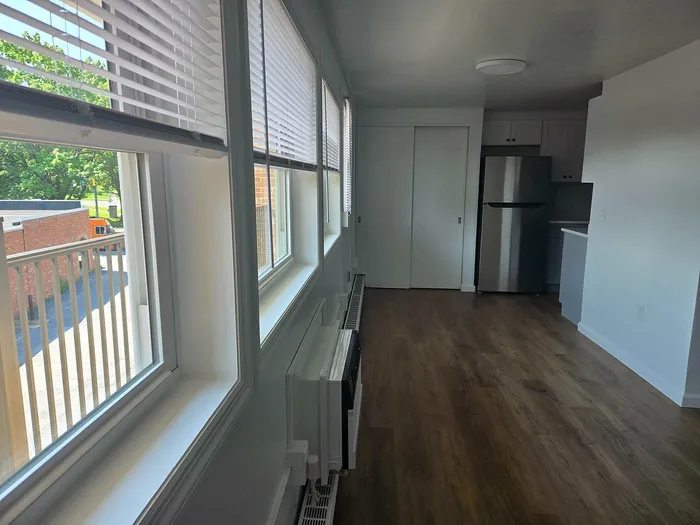 Bright, modern apartment with sleek wood flooring, large windows offering abundant natural light, and a stylish stainless steel refrigerator in an open kitchen.