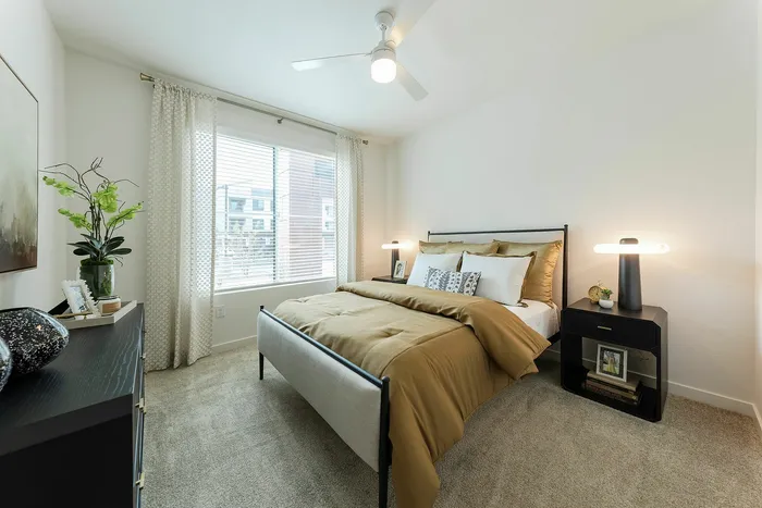 Two-Bedroom Senior Apartments In Phoenix, AZ - Liv+ Union Peak - Spacious Private Bedroom With Full-Sized Bed, Black Nightstand, Full Carpet, Large Windows, And A Dark Wood Dresser.