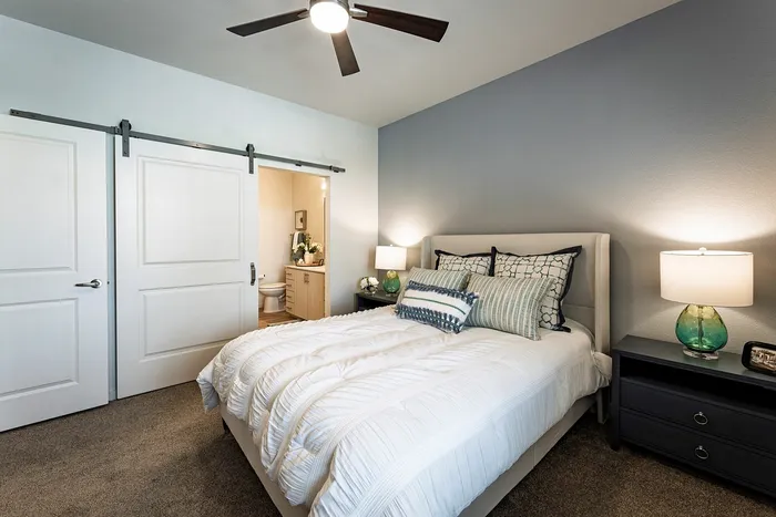 Elegant bedroom showcasing a cozy bed with plush pillows, modern ceiling fan, and stylish lighting, complemented by a sliding barn door leading to a sleek en-suite bathroom.