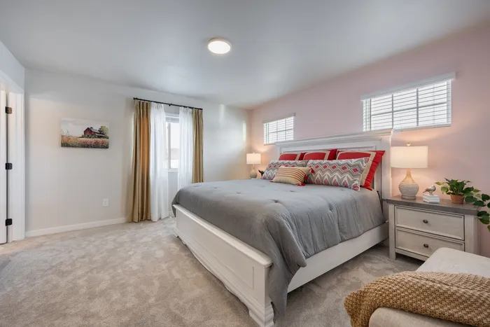 Inviting bedroom with soft pink walls, a cozy gray bed with stylish cushions, natural light from dual windows, and charming decor.