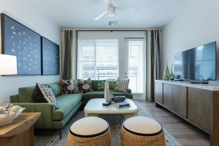 Modern living room featuring a plush green sectional sofa, elegant decor, and a flat-screen TV beneath large windows with city views.