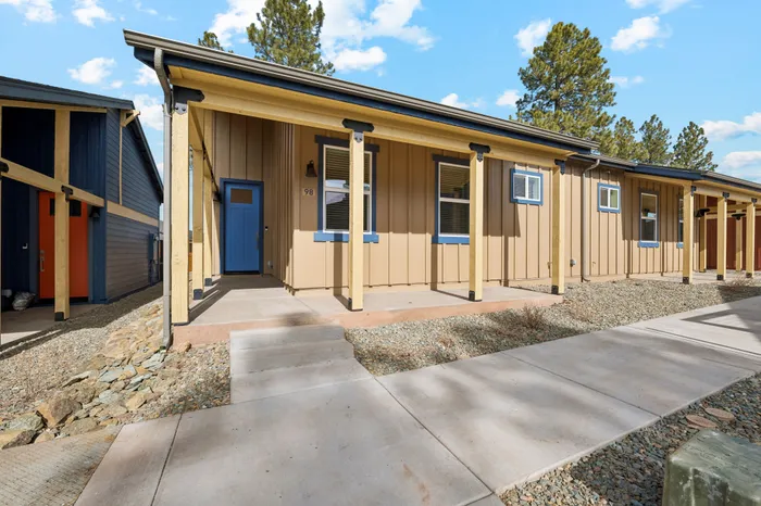 Charming single-story home with modern exterior, featuring a bright blue door and welcoming front porch, surrounded by scenic greenery and clear blue skies.