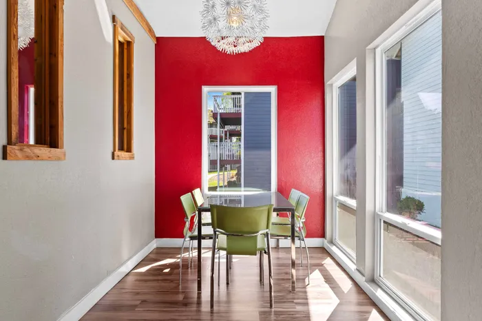Bright dining area with a modern chandelier, colorful red accent wall, large windows, and green chairs around a sleek table.