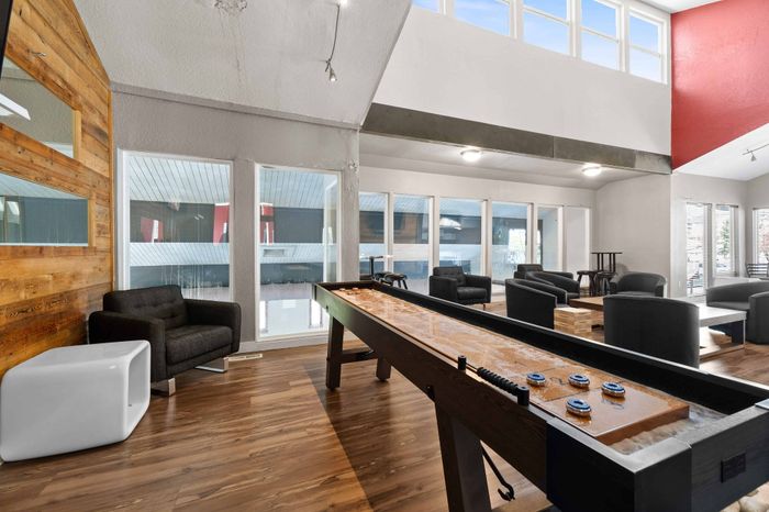 Bright and inviting community lounge featuring modern seating, a shuffleboard table, and expansive windows for natural light.