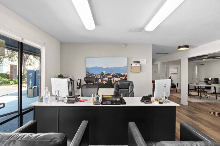 Inviting and modern office space featuring sleek black leather chairs, minimalist desks with computers, and a scenic landscape artwork, all bathed in natural light from large windows.