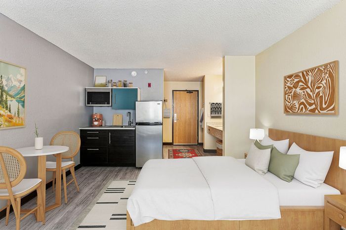 Modern studio apartment featuring a cozy bed with stylish decor, a compact kitchenette with stainless steel appliances, and a chic dining area with wooden chairs.
