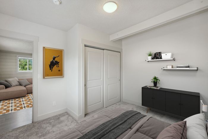 Inviting apartment bedroom featuring plush carpeting, a modern black sideboard, and wall-mounted shelves with stylish decor. Adjacent living room with cozy sofa visible through doorway.