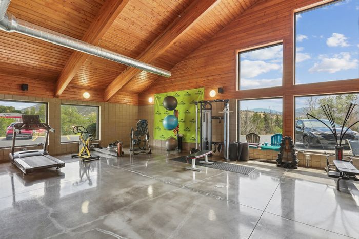 Spacious home gym with modern equipment, high wooden ceilings, and floor-to-ceiling windows offering scenic views.