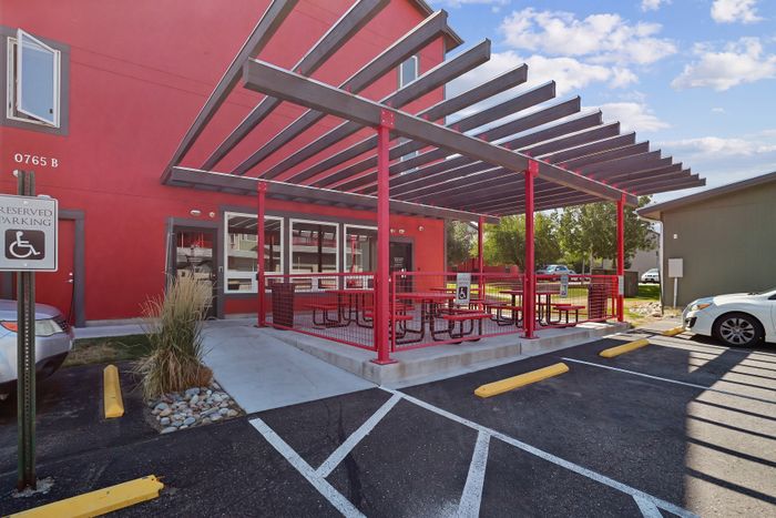 Modern exterior with a vibrant red fa&ccedil;ade and a stylish pergola-covered outdoor seating area, featuring accessible parking nearby.