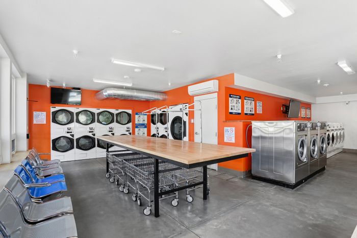 Modern laundry room featuring multiple washers and dryers, bright orange accents, and spacious folding area with handy carts.