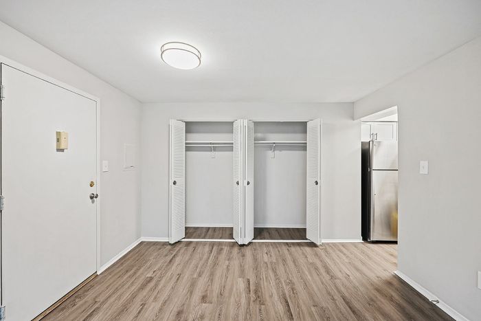 Spacious room with open double closets and sleek hardwood flooring, adjacent to a modern kitchen.