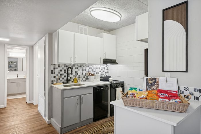 Bright apartment kitchen with sleek white cabinets, modern black appliances, and stylish checkered backsplash, complemented by a cozy wooden floor and a welcoming snack basket.