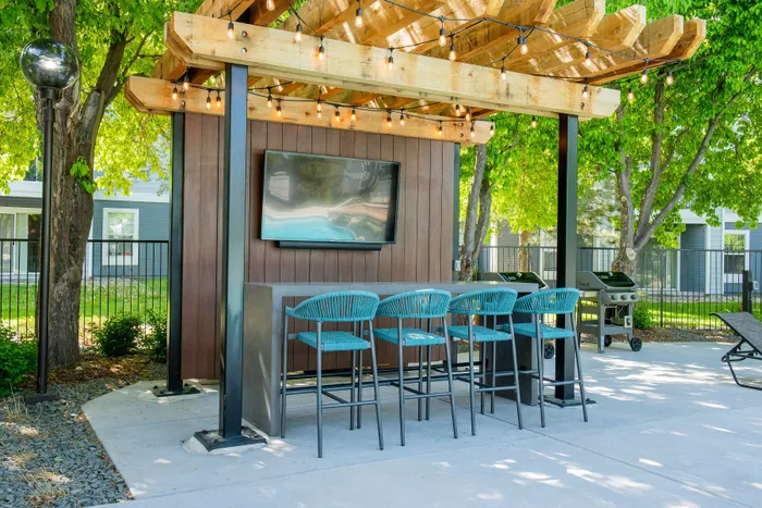 Charming outdoor lounge area featuring a flat-screen TV, modern bar seating, and bbq grill under a pergola with string lights, set against a lush green backdrop.