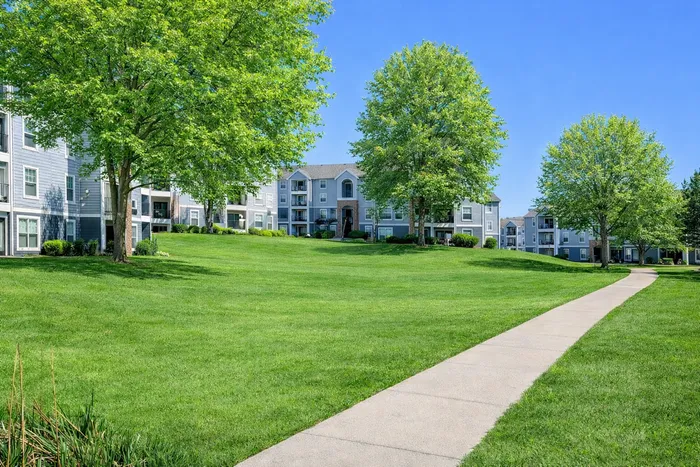 Bright and inviting apartment complex with lush green lawns, mature trees, and charming pathways under a clear blue sky.