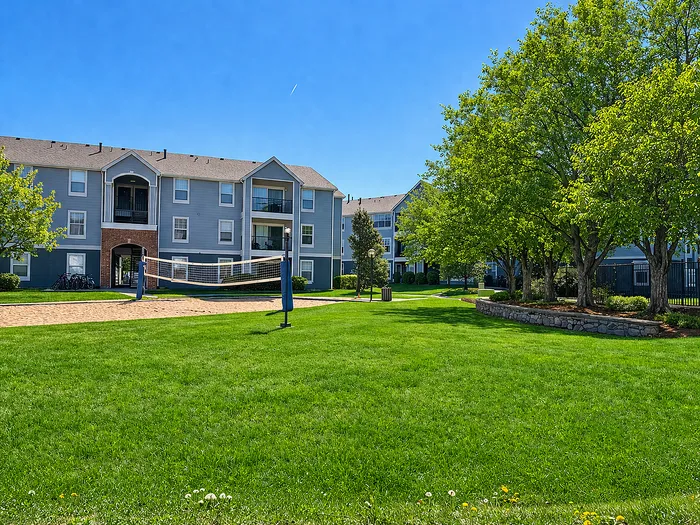 Charming apartment complex with modern design, surrounded by lush greenery and a vibrant outdoor volleyball court under a clear blue sky.