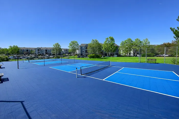 Beautiful apartment complex with vibrant blue tennis courts, lush green surroundings, and clear blue skies.