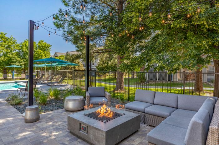 Charming outdoor patio with a cozy fire pit, modern seating, and a view of a sparkling pool surrounded by lush trees and ambient string lights.