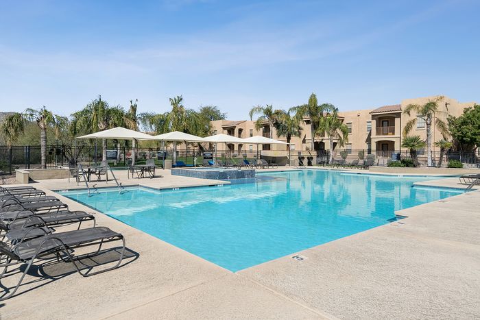 Luxurious apartment complex featuring a sparkling outdoor pool surrounded by elegant lounge chairs and shaded seating areas under pristine blue skies.
