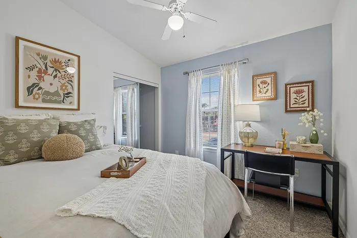 Cozy bedroom featuring chic decor with floral artwork, subtle blue accent wall, elegant desk space, and natural light shining through sheer curtains.