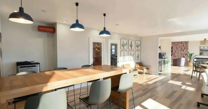 Modern, sunlit communal space featuring a large wooden table with stylish chairs, chic pendant lighting, and vibrant wall art, perfect for gatherings or co-working.