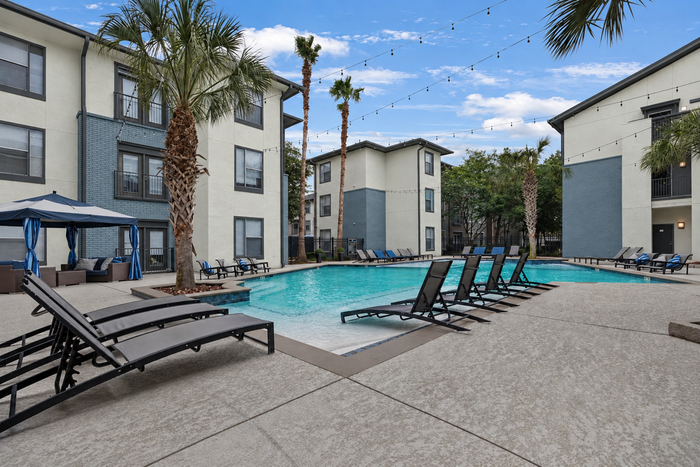 Elegant apartment community with a sparkling pool, surrounded by lounge chairs and shaded seating, enhanced by tall palm trees and modern architecture.
