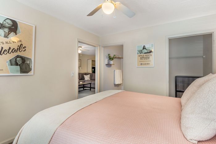 Bright bedroom with a soft pink bedspread, wall art, and view into a modern living area at Village Green in Raleigh, North Carolina.