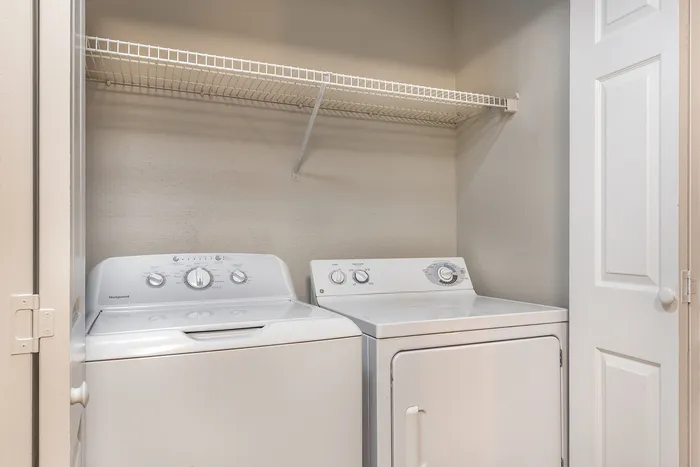 Compact laundry area featuring a washer and dryer with a convenient overhead storage shelf at Village Green in Raleigh, North Carolina..