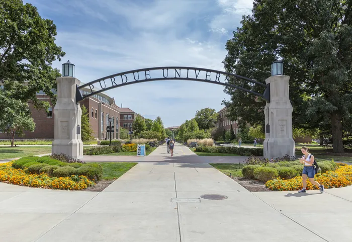 Iconic campus entrance with vibrant flowers and students strolling under a grand archway, creating a welcoming academic environment. West Lafayette, Indiana. Near Purdue.