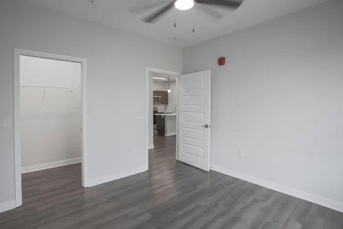 Bright bedroom with modern hardwood flooring, ceiling fan, and view into a sleek kitchen and spacious walk-in closet.