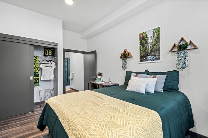 Cozy bedroom featuring a plush bed with green comforter and decorative pillows, stylish geometric wall shelves with plants, and an open closet displaying sports apparel.