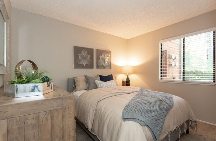 Cozy bedroom with earth-toned decor, featuring a plush bed, modern artwork, and natural light from a large window.