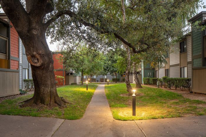 Charming apartment setting featuring a serene pathway lined with trees and soft lighting, nestled between modern, multi-story buildings.
