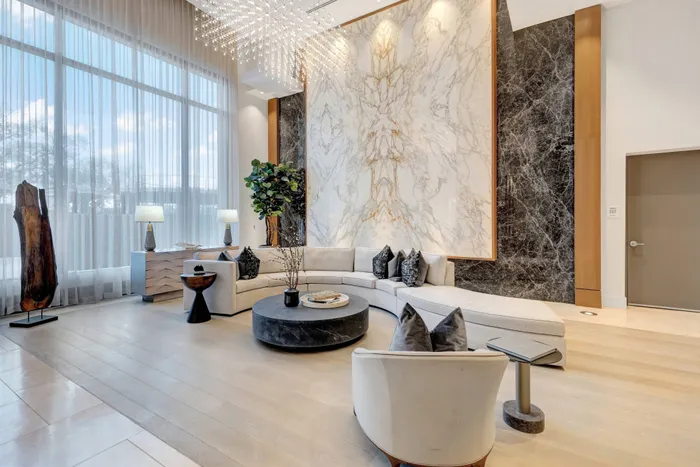 Spacious lobby with a long curved couch, high ceilings, full-width floor-to-ceiling windows, modern art, and a full-height book-matched marble focal piece on the wall.