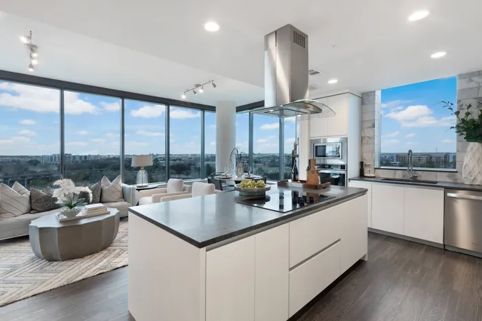 Open-concept living, dining and kitchen space with dark wood-style floors, a large island with an electric range and stainless steel overhead hood, white handleless cabinets, and a panoramic city view through two full-width floor-to-ceiling windows.