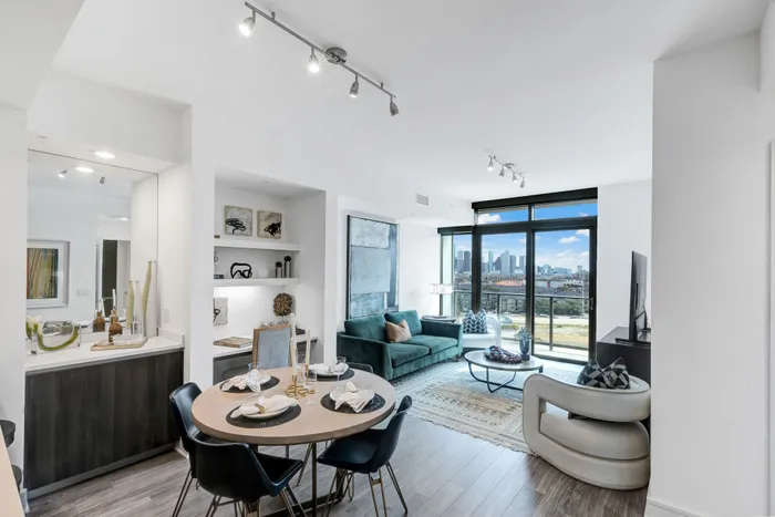 Open-concept living space with light ash-colored wood-style floors, floor-to-ceiling windows, a dry bar, dining table with 4 chairs, a built-in desk with shelves, and a sofa, coffee table, armchair, and media console in the living room.