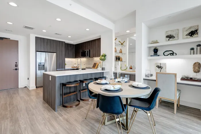 Open-concept kitchen and dining space with light ash-colored wood-style floors, dark cabinets, dry bar, dining table with 4 chairs, and a built-in desk with shelves and a chair.