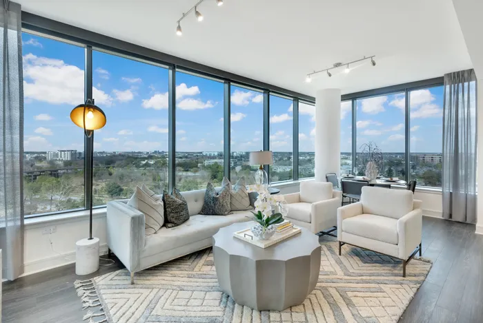 Bright living room with full-width windows on two walls up to the ceiling and a view of a park in the foreground and city in the background. The living room has a couch, two armchairs, a coffee table, and a dining area.