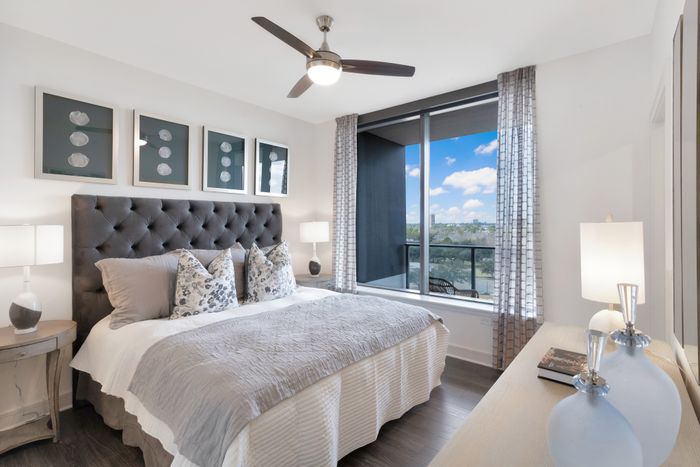 Bedroom with king-sized bed, dresser, white walls, dark wood-style floors, ceiling fan, and large window overlooking a balcony and a view of the park.