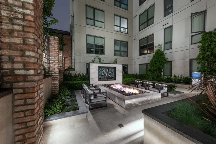 A landscaped stone courtyard with a large rectangular fire pit, 3 armchairs on each side, and an HDTV mounted on a freestanding stone wall.