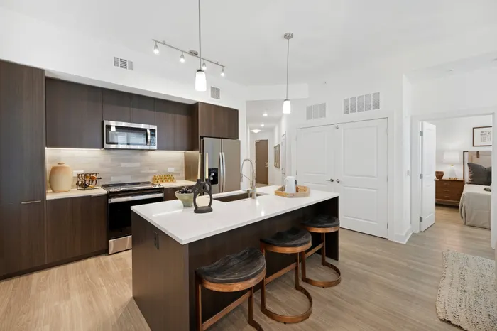 Kitchen with stainless-steel appliances, modern dark wood cabinetry, a white marble island with three bar stools and light wood-style floors.