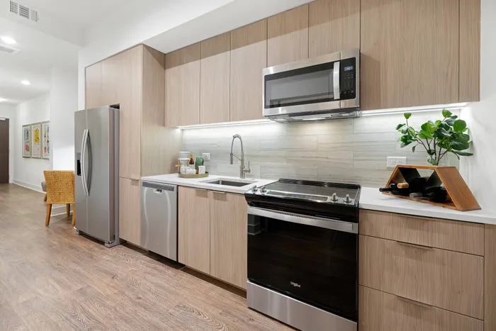 Studio apartment kitchen with stainless-steel appliances, modern light wood cabinetry, light wood-style floors and a tile backsplash.