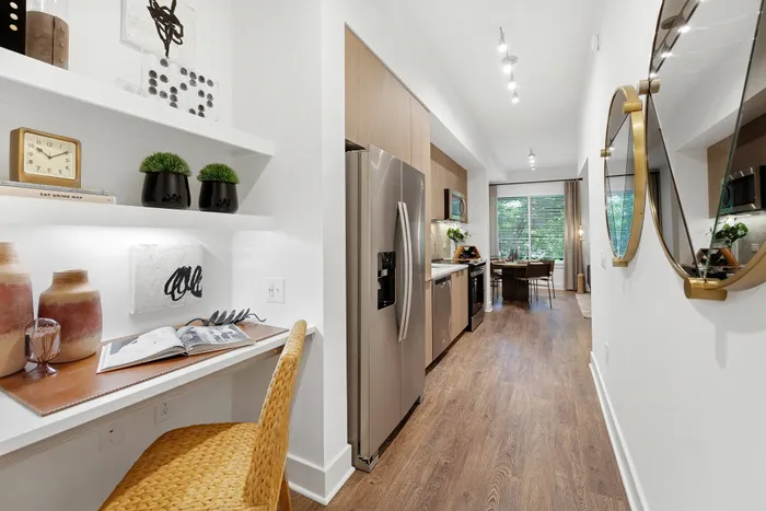 View from down the hall of a studio apartment kitchen with stainless-steel appliances. A built-in desk with shelves is in the foreground.