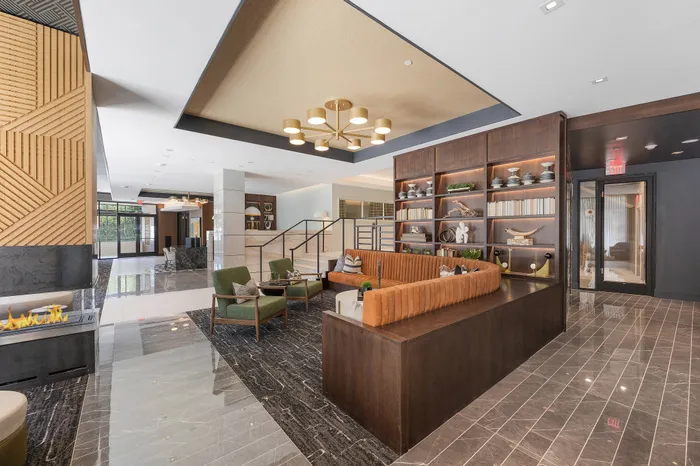 Spacious, modern lobby with an upholstered banquette, armchairs, a glass-walled fireplace, marble floors with gray carpeted areas, wood-paneled bookshelves and marble stairs.