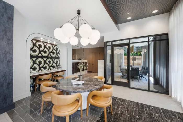 Four mid-century modern curved wood chairs around a gray marble table with a view into the glass-walled conference room. A desk area is set into the wall with a mosaic backsplash.